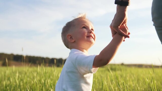 Walking hand in hand through grass child toddler holds parent hand while walking across sunlit field in summer nature setting family bond visible child smile gentle guiding hand soft sunlight