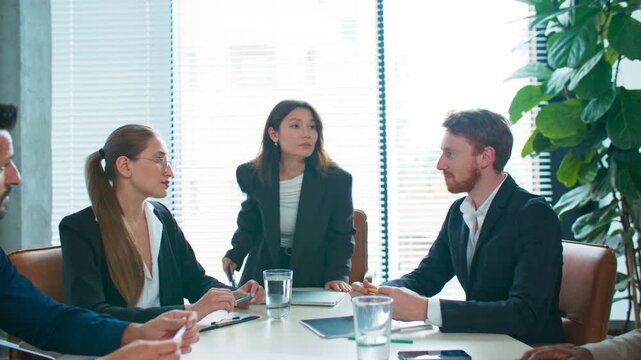 Professional woman in formal suit entering meeting and preparing talking with colleagues. Holding notebook with new data for discussion. Team waiting attentively. Starting conversation.