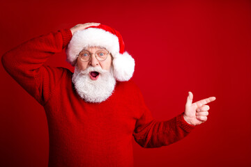 Joyful Santa in red sweater with white beard surprised and pointing against a bright red background