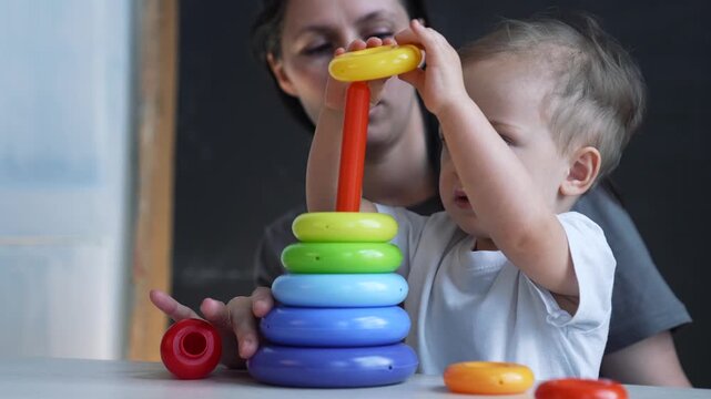 Toddler stacking colorful ring toy while mother guides hand, child focuses on ring placement, parent supports play and learning, promoting skill development and education through toy play now