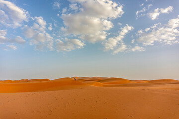 Wide landscape view of the golden sand dunes in the Erg Chebbi desert, Morocco, under a clear blue sky with scattered clouds. The calm and warm light highlights the textures of the desert surface.