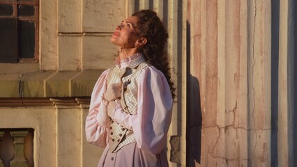 A woman dressed in an elegant historical dress stands near ancient columns, gently holding a letter to her chest in a moment of emotion and nostalgia under the warm rays of the sun.