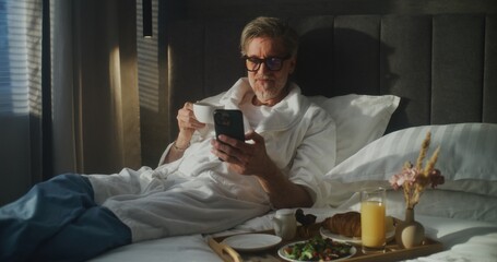 Morning Routine Coffee, Phone, and Comfort in Hotel Bed. Man in Bathrobe and Glasses Relaxes in Bed, Sipping Coffee While Scrolling on Smartphone. Business Travel Comfort, Digital Habits. Dolly Shot.