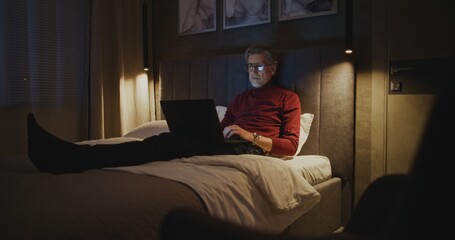 Mature Professional Sits on Bed Using Laptop Late at Night in Hotel Room. Man Types on Laptop Keyboard. Concept of Business Traveler Life, Modern Technology, and Late Night Productivity. Handheld.