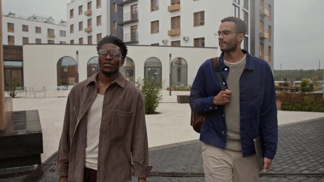 Two biracial colleagues carrying backpack and laptop, chatting and enjoying weather while walking in downtown