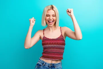 Fototapete Rund Musik Young woman celebrates success wearing a red patterned top against a blue background smiling and raising arms  © deagreez