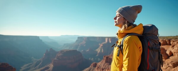 Young woman with backpack takes break on canyon edge enjoying view. Female hiker wears warm hat yellow jacket against blue sky. She breathes fresh air, feeling peacefulness, solitude.