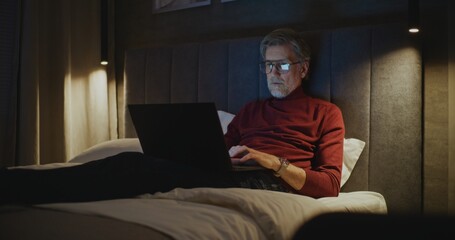 Elegant Mature Man Wearing Glasses Uses Laptop While Sitting On Bed. In Warm Evening Light and Calm Atmosphere, Man Works On His Personal Computer. Concept Travel, Digital Connectivity. Dolly Shot.