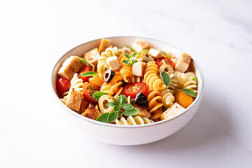 Warm salad with colorful full grain fusilli pasta, olives, fried tofu, cherry tomatoes and aromatic oregano leaves in ceramic bowl on white marble table top view. Healthy vegan food concept.