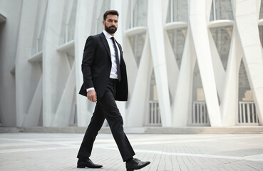Bearded man in elegant black suit strides confidently past modern building. Pro male walks with purpose, exuding success, determination. Business executive on city street, focused on career goals.