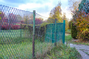 Green chain-link fence with metal poles bordering autumn park pathway in residential Warsaw neighborhood. High quality photograph.