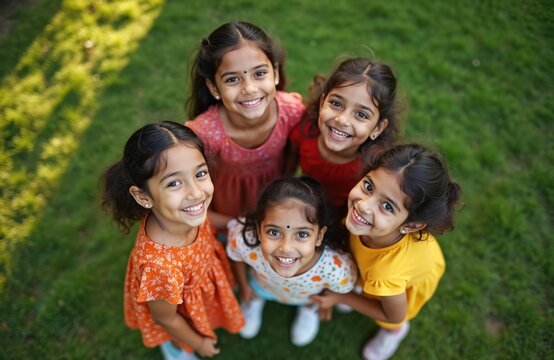 Five happy Indian children smile together in a grassy park looking up. Young girls form a circle outdoors, sharing a moment of joy and connection. Diverse childhood fun. - Powered by Adobe