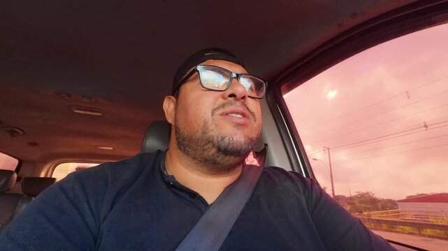 A plus-sized man wearing glasses and a cap drives a car while speaking, possibly vlogging or sharing thoughts during a road trip. 