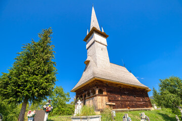 Church of the Archangels Michael and Gabriel, 18th century wooden spire rising over a wildflower strewn cemetery in Sisesti, Maramures, under a bright blue summer sky