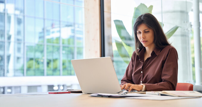 Young professional specialist latin hispanic business lady working on laptop pc at desk in modern office space. Middle eastern woman using computer technology app for work online. Banner, copy space