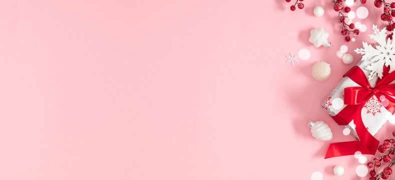 Festive Christmas composition with gift box, red ribbon, white snowflakes and berries on pink background. Modern and elegant holiday banner for winter celebrations.