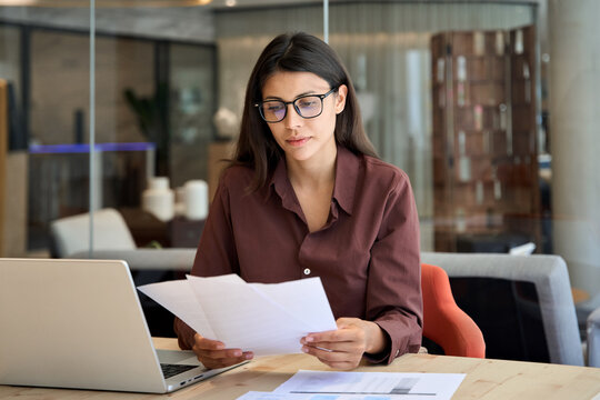 Focused latin hispanic business woman working on laptop computer reading financial document trader report in office. Thinking accountant entrepreneur manager businesswoman doing paperwork using pc 