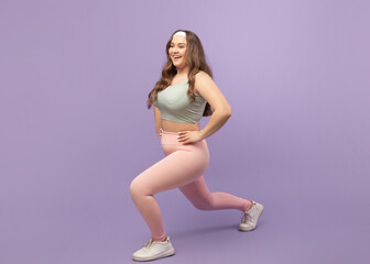 A young european plus size woman exercises with lunges on a purple background. She smiles while...