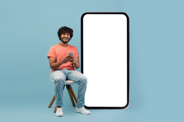 A cheerful young man with curly hair sits on a chair, smiling while using his cell phone. He is...