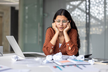 Young woman feeling overwhelmed and frustrated by her work, sitting at a desk with a laptop and crumpled papers, showing signs of stress and a tight deadline