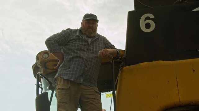 Medium shot of African American man as smiling farmer looking at camera and using digital tablet while standing in wheat field with combine harvester in background, copy space