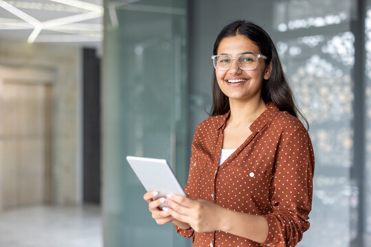 Young indian businesswoman in glasses and polka-dot shirt smiles confidently while holding a tablet in a bright office, conveying modern tech, leadership and professional success