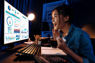 Smiling young beautiful asian creative looking on pc of profit of sales target report and writing on notebook for memo with planning invest new strategy market project at neon light night. Stratagem.