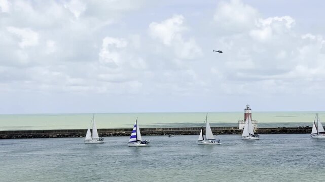 Helicopter flies over sailing regatta ensuring maritime safety as yachts leave port during sea festival.