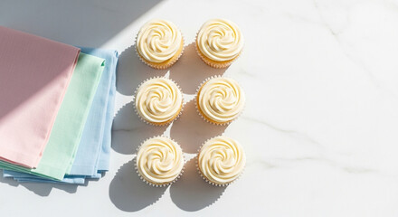 Six perfectly frosted vanilla cupcakes arranged elegantly on a white marble surface with pastel colored napkins nearby