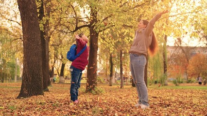 mother and little child in an autumn park throw dry leaves up, happy family, live fun with mom, cheerful kid plays with foliage and parent hands throwing leaf fall, parental care of girl, nature walk.