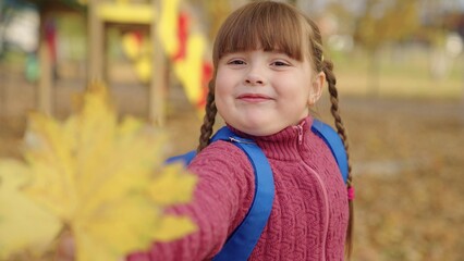 little girl with a backpack on her shoulders holds yellow leaves in her hand and smiles, golden...
