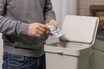 Man discarding cash into a trash bin, symbolizing wastefulness and financial irresponsibility in a domestic setting with a modern kitchen backdrop
