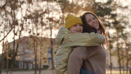 little child is having fun with her mother in city park, happy family life, kid girl on her back mom laughs, childish desires play, daughter love hug mom, laughing cheerful people on street glare sun.