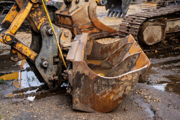 Close-up of old excavator bucket attached to hydraulic arm at muddy construction site showing rust, wear and mechanical detail of heavy machinery used for earthmoving and industrial groundworks