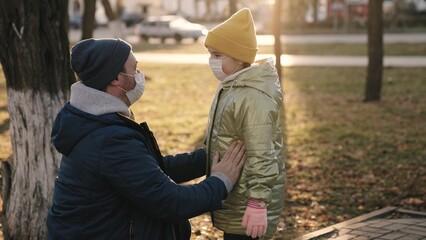 Father adjusts protective medical mask on face of little daughter in orange hat on blurred background during pandemic close view. Happy family. Child with dad on playground. Children walk with daddy