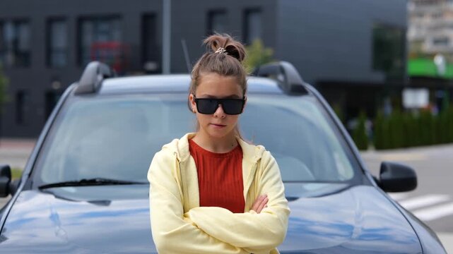 Pretty cool young teenager girl in black sunglasses standing in front of car on street looking at camera chewing a gum. Close up. Stylish pretentious teen smiling at camera.