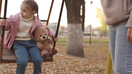 a little child sways with a teddy bear in an autumn park, a happy kid dreams of flying with his favorite toy, an active family vacation of mother and daughter on walk in city, childhood friend of girl