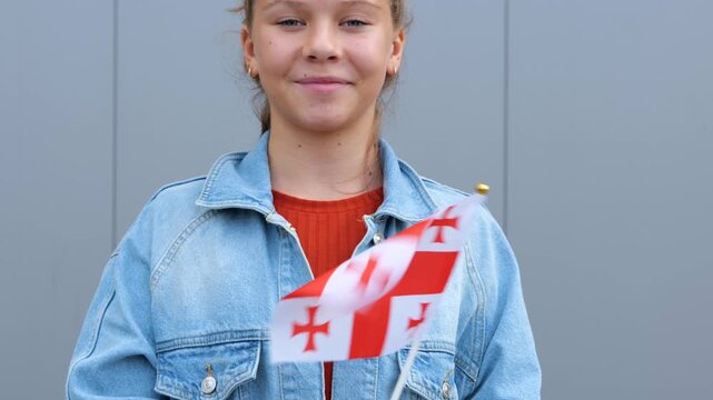 Young female is standing outdoors, smiling and waving small Georgian flag. Caucasian beautiful teenage girl is keeping small flag of Georgia in her hands.