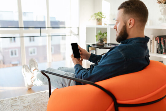 A man relaxes in a vibrant orange chair, focused on his smartphone. Sunlight streams through large windows, highlighting the modern decor of the room.