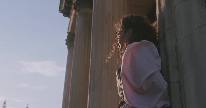 Close up of a woman in historical clothing, looking pensively next to ancient stone columns, illuminated by soft golden evening light.