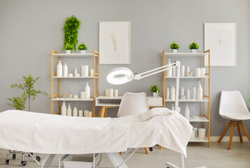 Clean and bright spa treatment room with an empty massage bed, towel, magnifying lamp, shelves of...