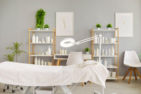 Clean and bright spa treatment room with an empty massage bed, towel, magnifying lamp, shelves of cosmetic products. The image represents a modern, professional, and relaxing beauty care environment.