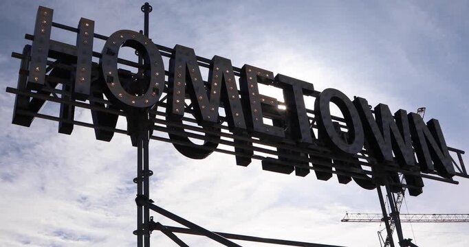 Big metal marquee letters spelling HOMETOWN rise against a bright sky. Warm bulbs and a sun flare suggest belonging, roots, civic pride, and the idea of home in an urban landscape.