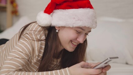 cheerful girl sends a message to the chat and laughs, merry christmas greetings, a teenager in a santa hat smiles looking into a smartphone, a happy family, a teenager's childhood dream, new year mood