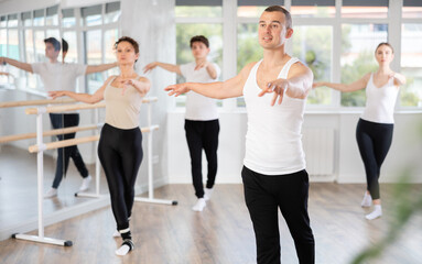 Fototapeta premium Sporty and graceful ballet artist in white tank top and black athletic pants confidently leading group rehearsal with amateur dancers, mirroring his movements in spacious serene dance studio