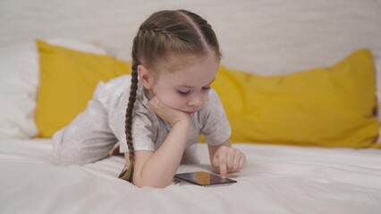 little child plays fun phone game lying on bed in room, a happy kid learns through smartphone application, children's education online through gadget sensor, girl daughter does homework with pleasure.