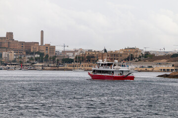 Fototapeta premium A red and white boat sails on the water in front of a city with historical buildings and a tall tower under an overcast sky.