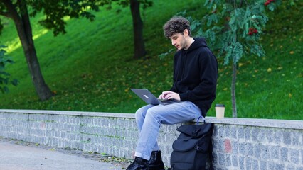Fototapeta premium Young man sitting in the park and typing on the laptop, coffee next to him. Autumn season. Education, working concept. Slow motion