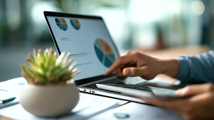 A middle aged man analyzes charts on a laptop a woman swiping through data on a tablet their desk busy with documents and a succulent plant top view financial analysis - Powered by Adobe