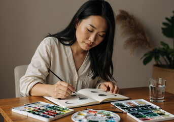 Fototapeta premium young asian woman painting in sketchbook at home with watercolors and paintbrush